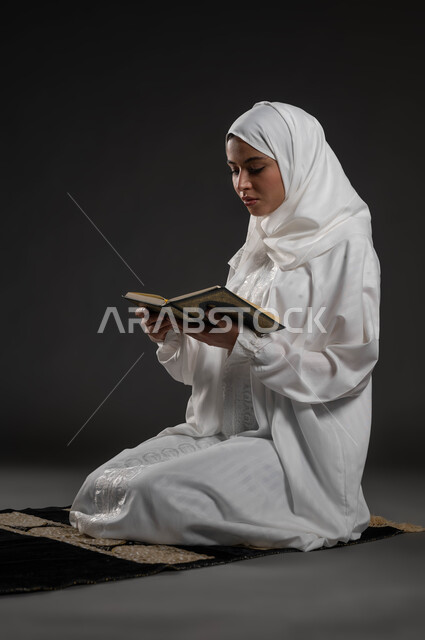 Recitation and reading of the Book of God Almighty, diligence and reverence in worship, portrait of a Saudi Gulf Arab woman wearing the hijab and white abaya, holding the Holy Qur’an in her hand, sitting on the prayer rug, getting closer to the Lord of the Worlds in the blessed month of Ramadan, gray background