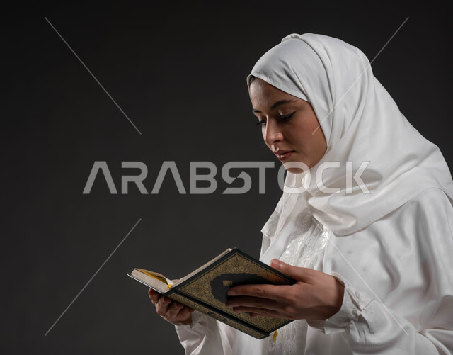 تلاوة وقراءة كتاب الله , إيماءات الخشوع و الاجتهاد في العباده, التقرب الى رب العالمين في شهر رمضان المبارك،  بورتريه مقرب امرأة عربيه خليجية سعودية ترتدي الحجاب و العباءة البيضاء وتمسك بيدها القرآن الكريم، خلفية رمادية
