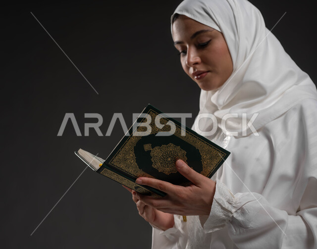 إيماءات الخشوع و الاجتهاد في العباده, التقرب الى رب العالمين في شهر رمضان المبارك،  بورتريه مقرب امرأة عربيه خليجية سعودية ترتدي الحجاب و العباءة البيضاء وتمسك بيدها القرآن الكريم تقوم بالقراءة، خلفية رمادية