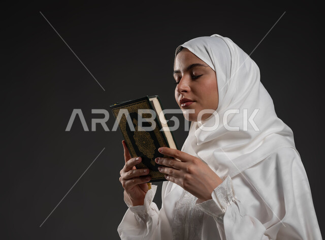 Closing the eyes to feel reassured and, insisting on supplication and requesting from God Almighty, worship and getting closer to the Lord of the worlds in the blessed month of Ramadan, close-up portrait of a Saudi Gulf Arab woman wearing the hijab and white abaya and holding the Holy Quran in her hand, gray background.
