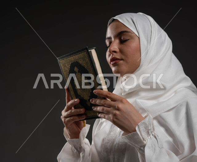 Insistence in supplication and request from God Almighty, worship and closeness to the Lord of the Worlds in the blessed month of Ramadan, gestures of reverence and reassurance, close-up portrait of a Saudi Gulf Arab woman wearing the hijab and white abaya, closing her eyes, holding the Holy Qur’an in her hand, gray background
