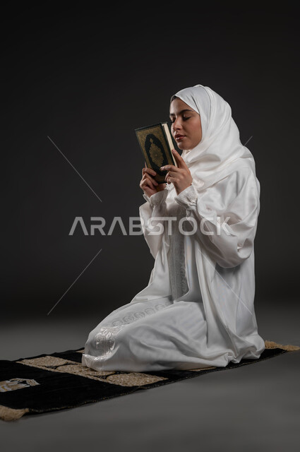 الإلحاح في الدعاء والطلب من الله تعالى, العبادة والتقرب الى رب العالمين في شهر رمضان المبارك، إيماءات الخشوع و الطمأنينة, بورتريه لامرأة عربيه خليجية سعودية ترتدي الحجاب و العباءة البيضاء تمسك بيدها القرآن الكريم تجلس على سجادة الصلاه, خلفية رمادية
