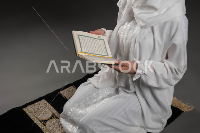 قراءة كتاب الله تعالى, إيماءات الخشوع و الإجتهاد في العباده, بورتريه مقرب لأيدي امرأة عربية سعودية خليجية ترتدي الحجاب و العباءة البيضاء تمسك بيدها القرآن الكريم تجلس على سجادة الصلاه, التقرب الى رب العالمين في شهر رمضان المبارك، خلفية رمادية