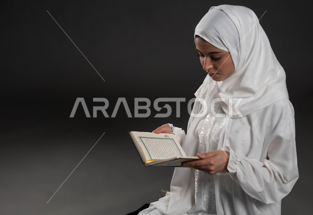 التقرب الى رب العالمين في شهر رمضان المبارك، الإجتهاد و الخشوع في العباده, بورتريه لامرأة عربية سعودية خليجية ترتدي الحجاب و العباءة البيضاء تمسك بيدها القرآن الكريم ،قراءة كتاب الله تعالى, خلفية رمادية