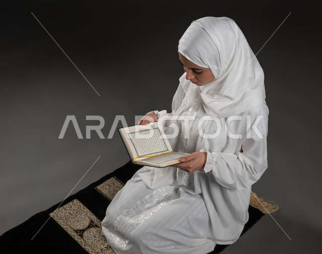 الإجتهاد و الخشوع في العباده, بورتريه لامرأة عربية سعودية خليجية ترتدي الحجاب و العباءة البيضاء تمسك بيدها القرآن الكريم تجلس على سجادة الصلاه, قراءة كتاب الله تعالى, التقرب الى رب العالمين في شهر رمضان المبارك، خلفية رمادية