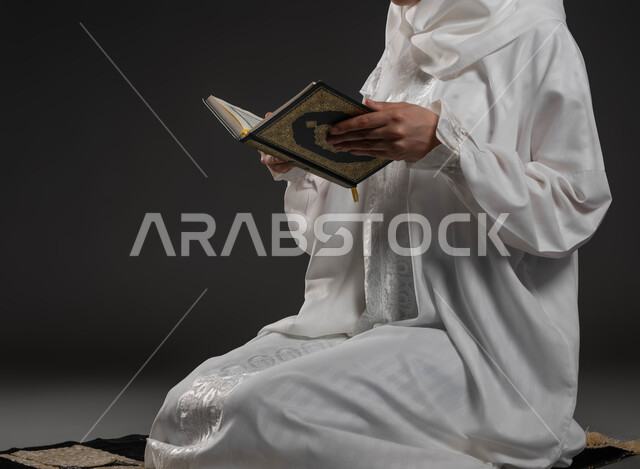 قراءة كتاب الله تعالى, إيماءات الخشوع و الإجتهاد في العباده, بورتريه مقرب لأيدي شابة عربية خليجية سعودية ترتدي العباءة البيضاء تمسك بيدها القرآن الكريم تجلس على سجادة الصلاه تقوم بالتلاوة, التقرب الى رب العالمين في شهر رمضان المبارك، خلفية رمادية
