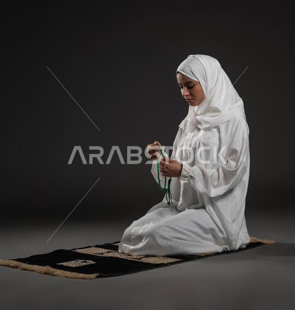قراءة الأدعية والأذكار، التسبيح والتهليل وذكر الله تعالى، بورتريه لسيدة عربية خليجية سعودية محجبة ترتدي العباءة البيضاء تجلس على سجادة الصلاة تمسك بيدها السبحة تجلس على السجادة، العبادة والتقرب لرب العالمين، خلفية رمادية