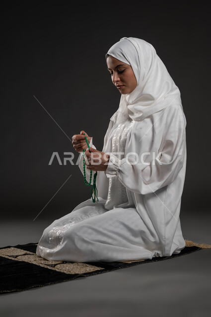 قراءة الأذكار والأدعية، التسبيح والتهليل وذكر الله تعالى، بورتريه لسيدة عربية خليجية سعودية محجبة ترتدي العباءة البيضاء تجلس على سجادة الصلاة تمسك بيدها السبحة تجلس على السجادة، العبادة والتقرب لرب العالمين، خلفية رمادية