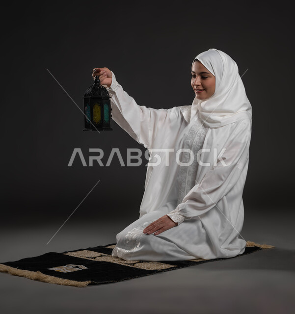 الاحتفال بقدوم شهر رمضان المبارك ، الأعياد والمناسبات الدينية، أجواء السكينة والروحانية، بورتريه لسيدة عربية خليجية سعودية محجبة ترتدي العباءة البيضاء تجلس على سجادة الصلاة تحمل بيدها فانوس معدني تنظر إليه، العبادة والتقرب إلى الله، خلفية رمادية