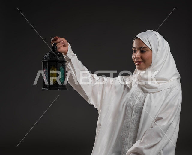 الأعياد والمناسبات الدينية، أجواء السكينة والروحانية، بورتريه لسيدة عربية خليجية سعودية محجبة ترتدي العباءة البيضاء تحمل بيدها فانوس معدني تنظر إليه، الإستعداد لقدوم شهر رمضان المبارك ، العبادة والتقرب إلى الله، خلفية رمادية