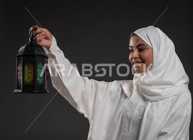 الأعياد والمناسبات الدينية، أجواء السكينة والروحانية، بورتريه لسيدة عربية خليجية سعودية محجبة ترتدي العباءة البيضاء تحمل بيدها فانوس معدني تنظر إليه، الإستعداد لقدوم شهر رمضان المبارك ، العبادة والتقرب إلى الله، خلفية رمادية