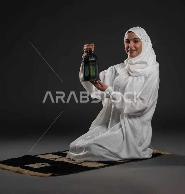 المناسبات والأعياد الدينية، أجواء السكينة والروحانية، بورتريه لسيدة عربية خليجية سعودية محجبة ترتدي العباءة البيضاء تنظر إلى الكاميرا تجلس على سجادة الصلاة تحمل بيدها فانوس معدني، الإستعداد لقدوم شهر رمضان المبارك ، العبادة والتقرب إلى الله، خلفية رمادية
