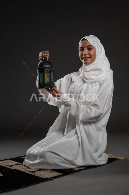 أجواء السكينة والروحانية، بورتريه لسيدة عربية خليجية سعودية محجبة ترتدي العباءة البيضاء تنظر إلى الكاميرا تجلس على سجادة الصلاة تحمل بيدها فانوس معدني، الإستعداد لقدوم شهر رمضان المبارك ، المناسبات والأعياد الدينية، العبادة والتقرب إلى الله، خلفية رمادية