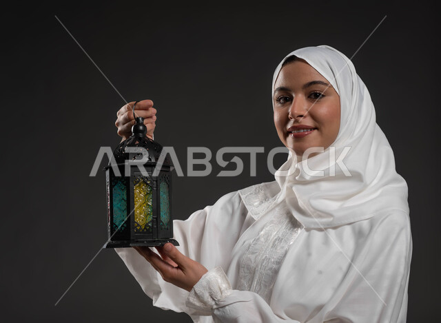 الإستعداد لقدوم شهر رمضان المبارك، أجواء السكينة والروحانية، بورتريه مقرب لسيدة عربية خليجية سعودية محجبة ترتدي العباءة البيضاء تنظر إلى الكاميرا تحمل بيدها فانوس معدني، المناسبات والأعياد الدينية، العبادة والتقرب إلى الله، خلفية رمادية