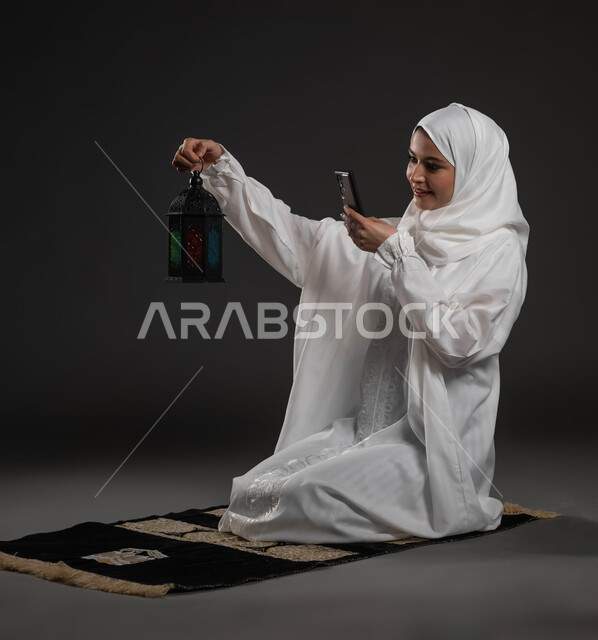 الأجواء الروحانية في شهر رمضان المبارك، بورتريه لإمرأة عربية سعودية خليجية ترتدي الحجاب والعباءة البيضاء تجلس على سجادة الصلاة تمسك بيدها الهاتف المحمول تقوم بالتقاط صورة لفانوس معدني، العبادة والتقرب إلى الله، خلفية رمادية