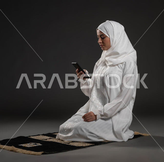 مفهوم التطبيقات والبرامج الدينية، قراءة الأذكار والأدعية، بورتريه مقرب لإمرأة عربية سعودية خليجية ترتدي الحجاب والعباءة البيضاء تجلس على سجادة الصلاة تمسك بيدها الهاتف المحمول تقوم بتلاوة القرآن من المصحف الالكتروني عبر تطبيقات الجوال، خلفية رمادية