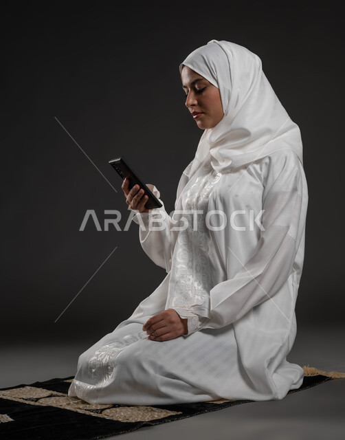 مفهوم البرامج والتطبيقات الدينية، قراءة الأذكار والأدعية، بورتريه مقرب لإمرأة عربية سعودية خليجية ترتدي الحجاب والعباءة البيضاء تجلس على سجادة الصلاة تمسك بيدها الهاتف المحمول تقوم بتلاوة القرآن من المصحف الالكتروني عبر تطبيقات الجوال، خلفية رمادية