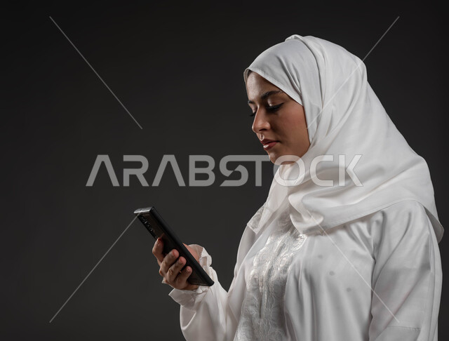 قراءة الأذكار والأدعية، بورتريه مقرب لإمرأة عربية سعودية خليجية ترتدي الحجاب والعباءة البيضاء تمسك بيدها الهاتف المحمول تقوم بتلاوة القرآن من المصحف الالكتروني عبر تطبيقات الجوال، العبادة والتقرب إلى الله، خلفية رمادية
