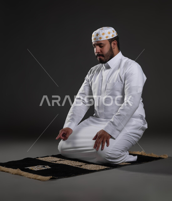 العبادة والتقرب إلى رب العالمين، الخشوع في الصلاة والتضرع الى الله،  بورتريه لرجل عربي خليجي سعودي يرتدي الثوب والقبعة الإسلامية البيضاء يجلس على السجادة يقوم بالتشهد، آداء الفرائض والعبادات، خلفية رمادية
