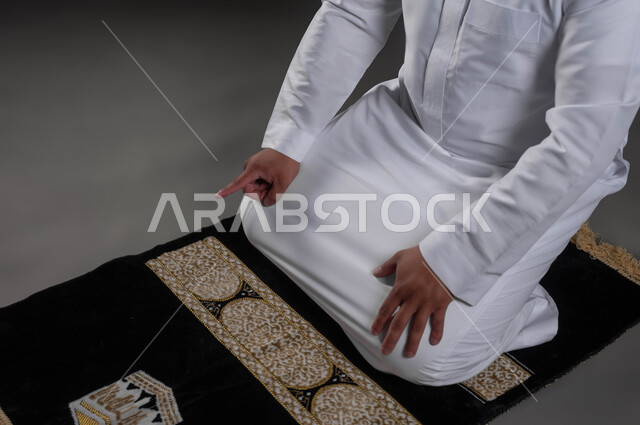 الشعور بالسكينة والطمأنينة، الخشوع في الصلاة والتضرع الى الله، بورتريه مقرب لأيدي رجل عربي خليجي سعودي يرتدي الثوب الابيض يجلس على السجادة يرفع إصبع السبابة للتشهد، آداء الفرائض والعبادات، خلفية رمادية