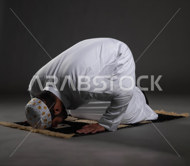 آداء الفرائض والعبادات، الخشوع في الصلاة والتضرع الى الله، بورتريه مقرب لرجل عربي خليجي سعودي يرتدي الثوب والقبعة الإسلامية البيضاء يقوم بالسجود، الشعور بالسكينة والطمأنينة، خلفية رمادية