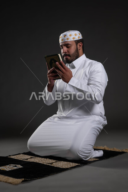 الإكثار من الدعاء والطلب من الله تعالى, العبادة والتقرب الى رب العالمين، إيماءات الخشوع و الطمأنينة, بورتريه لرجل عربي سعودي خليجي يرتدي الثوب والقبعة الإسلامية البيضاء يغمض عينيه يمسك بيده القرآن الكريم يجلس على سجادة الصلاة, خلفية رمادية