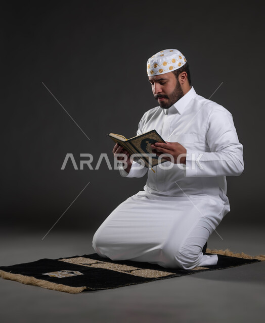 العبادة والتقرب الى رب العالمين في شهر رمضان المبارك، قراءة كتاب الله تعالى, إيماءات الخشوع و الطمأنينة, بورتريه لرجل عربي سعودي خليجي يرتدي الثوب والقبعة الإسلامية البيضاء يمسك بيده القرآن الكريم يجلس على سجادة الصلاه, خلفية رمادية