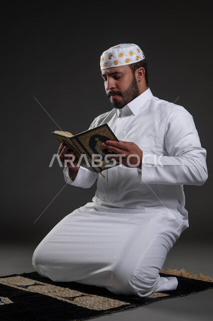 قراءة كتاب الله تعالى, إيماءات الخشوع و الإجتهاد في العباده, بورتريه لرجل عربي سعودي خليجي يرتدي الثوب والقبعة الإسلامية البيضاء يمسك بيده القرآن الكريم يجلس على سجادة الصلاه, التقرب الى رب العالمين في شهر رمضان المبارك، خلفية رمادية