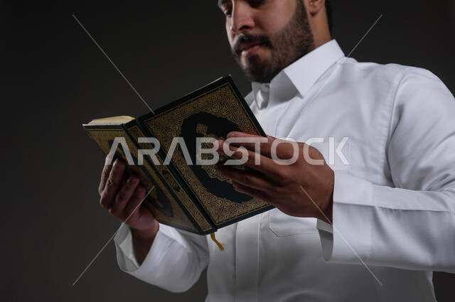 التقرب الى رب العالمين في شهر رمضان المبارك، إيماءات الخشوع و الإجتهاد في العباده, بورتريه مقرب لرجل عربي سعودي خليجي يرتدي الثوب الأبيض يمسك بيده القرآن الكريم يقوم بالقراءة، خلفية رمادية