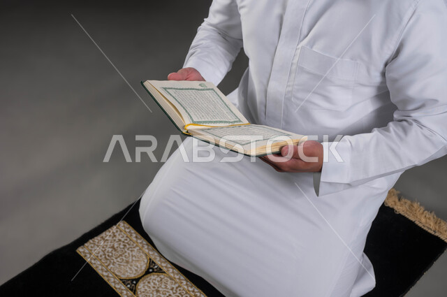 قراءة كتاب الله تعالى, إيماءات الخشوع و الإجتهاد في العباده, بورتريه مقرب لأيدي رجل عربي سعودي خليجي يرتدي الثوب والقبعة الإسلامية البيضاء يمسك بيده القرآن الكريم يجلس على سجادة الصلاه, التقرب الى رب العالمين في شهر رمضان المبارك، خلفية رمادية