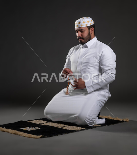 قراءة الأدعية والأذكار، التسبيح والتهليل وذكر الله تعالى، بورتريه لرجل عربي خليجي سعودي يرتدي الثوب والقبعة الإسلامية البيضاء يمسك بيده السبحة يجلس على السجادة، العبادة والتقرب لرب العالمين، خلفية رمادية
