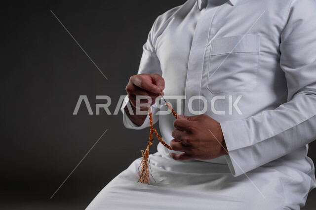 التهليل والتسبيح وذكر الله تعالى، بورتريه مقرب لأيدي رجل عربي خليجي سعودي يرتدي الثوب الأبيض يمسك بيده السبحة يجلس على السجادة، قراءة الأدعية والأذكار، مناسبات دينية وإسلامية، العبادة والتقرب لرب العالمين، خلفية رمادية