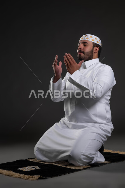 الدعاء و التضرع لرب العباد، أداء فرائض الصلاة بأوقاتها، مفهوم أركان الإسلام، بورتريه لرجل عربي خليجي سعودي يرتدي الثوب والقبعة الإسلامية البيضاء يجلس على السجادة، شاب وسيم مسلم، العبادة والتقرب إلى الله، صورة لكامل الجسم، خلفية رمادية