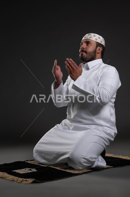 Supplication at the end of the prayer, performing the obligatory prayers at their times, the concept of the pillars of Islam, a portrait of a Saudi Gulf Arab man wearing the white Islamic dress and hat sitting on the carpet, a handsome young Muslim man, worship and getting closer to God, a full-body portrait, gray background