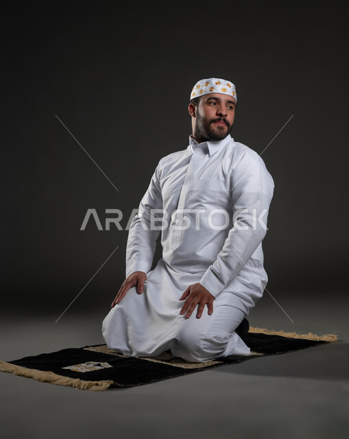 الشعور بالسكينة والطمأنينة، الخشوع والتضرع الى الله، بورتريه لرجل عربي خليجي سعودي يرتدي الثوب والقبعة الإسلامية البيضاء يجلس على السجادة يقوم بالسلام لإنهاء الصلاة، آداء الفرائض والعبادات، مفهوم أركان الإسلام، صورة لكامل الجسم، خلفية رمادية