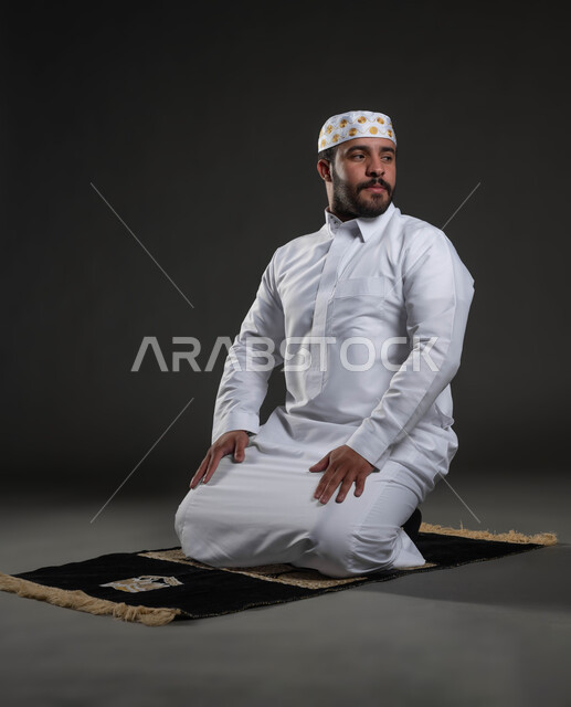 أداء الفرائض والعبادات، الخشوع و التضرع لله، الشعور بالسكينة والطمأنينة، بورتريه لرجل عربي خليجي سعودي يرتدي الثوب والقبعة الإسلامية البيضاء يجلس على السجادة يقوم بالسلام لإنهاء الصلاة، مفهوم أركان الإسلام، صورة لكامل الجسم، خلفية رمادية