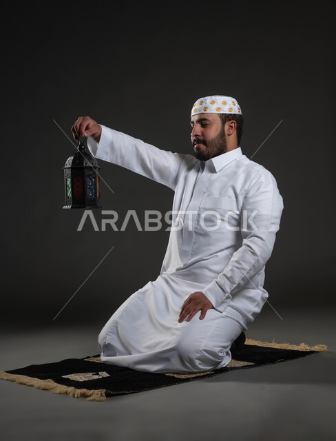 النظر لفانوس رمضان بإيماءات السعادة، العبادة والتقرب إلى الله، الاستمتاع بالأجواء الروحانية في لبشهر المبارك، بورتريه لرجل عربي سعودي خليجي يرتدي الثوب والقبعة البيضاء  يجلس على سجادة الصلاة، صورة لكامل الجسم، خلفية رمادية