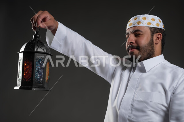 إيماءات السعادة والسرور، النظرإلى فانوس رمضان، العبادة والتقرب إلى الله، الاستمتاع بالأجواء الروحانية في لبشهر المبارك، بورتريه مقرب لرجل عربي سعودي خليجي يرتدي الثوب والقبعة البيضاء  يجلس على سجادة الصلاة، خلفية رمادية