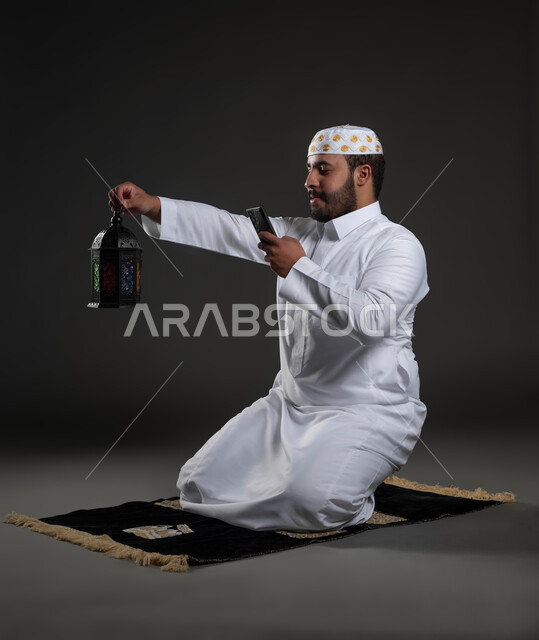 التقاط الصور لفانوس رمضان بإيماءات السعادة، العبادة والتقرب إلى الله، الاستمتاع بالأجواء الروحانية في لبشهر المبارك، بورتريه لرجل عربي سعودي خليجي يرتدي الثوب والقبعة البيضاء  يجلس على سجادة الصلاة يمسك بيده الهاتف المحمول، صورة لكامل الجسم، خلفية رمادية