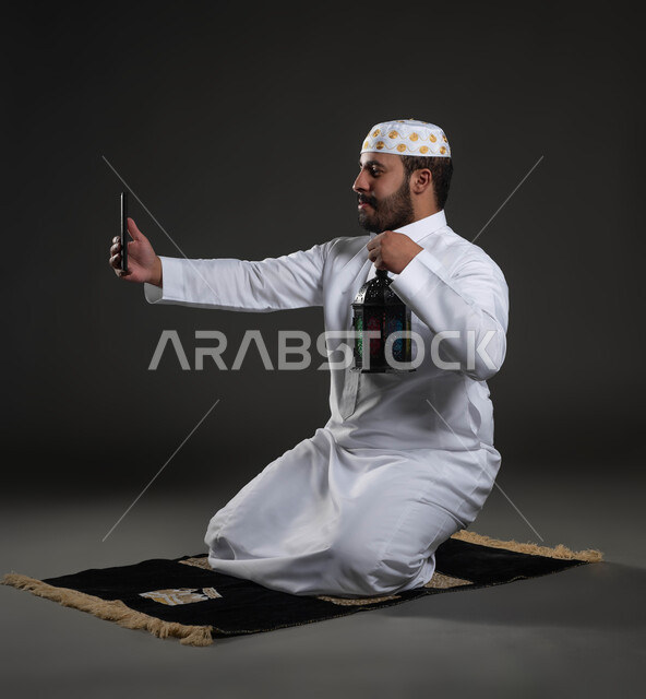 التقاط الصور لفانوس رمضان، العبادة والتقرب إلى الله، الاستمتاع بالأجواء الروحانية في لبشهر المبارك، بورتريه لرجل عربي سعودي خليجي يرتدي الثوب والقبعة البيضاء  يجلس على سجادة الصلاة يمسك بيده الهاتف المحمول، صورة لكامل الجسم، خلفية رمادية
