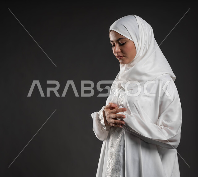 قراءة ما تيسر من القرآن الكريم، الركوع و السجود والخشوع، مفهوم أركان الإسلام ،أداء فرائض الصلاة بأوقاتها، بورتريه مقربلإمرأة عربية خليجية سعودية مسلمة ترتدي الحجاب والعباءة البيضاء تقف على السجادة، العبادة والتقرب إلى الله، خلفية رمادية