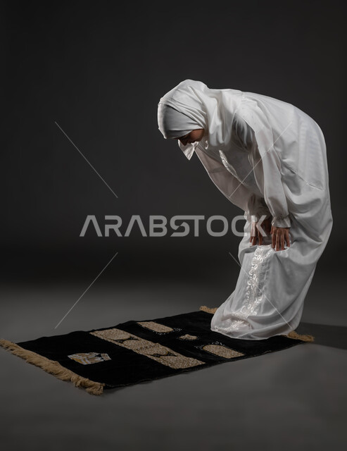 الركوع و السجود والخشوع، مفهوم أركان الإسلام ،أداء فرائض الصلاة بأوقاتها، بورتريه لإمرأة عربية خليجية سعودية مسلمة ترتدي الحجاب والعباءة البيضاء تقف على السجادة، العبادة والتقرب إلى الله، قراءة ما تيسر من القرآن الكريم، صورة لكامل الجسم،خلفية رمادية
