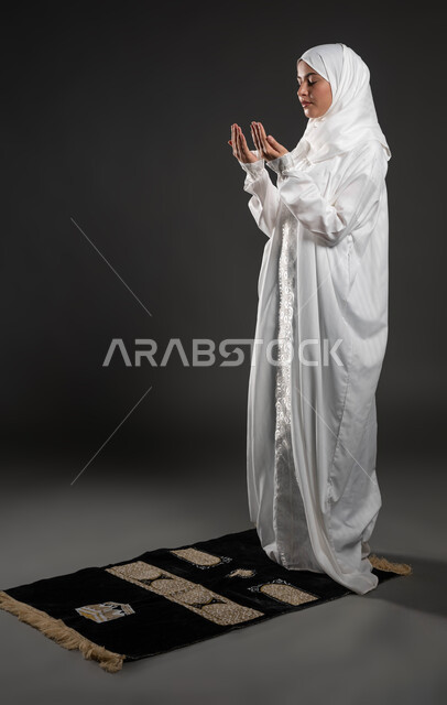 The concept of the pillars of Islam, performing the obligatory duties on time, supplication and supplication to the Lord of servants, worship and drawing closer to God, kneeling, prostrating, and humility, a close-up portrait of a young Saudi Gulf Arab Muslim woman wearing the hijab and white abaya, standing on the carpet, gray background.