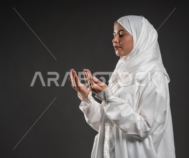 الدعاء والتضرع لرب العباد، العبادة والتقرب إلى الله، الركوع والسجود والخشوع، أداء الفرائض بأوقاتها، مفهوم أركان الإسلام، بورتريه مقرب لشابة عربية خليجية سعودية مسلمة ترتدي الحجاب و العباءة البيضاء تقف على السجادة، خلفية رمادية