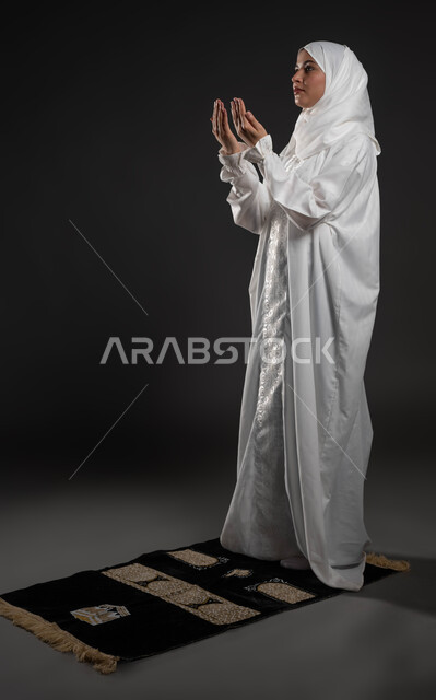 العبادة والتقرب إلى الله، الركوع والسجود والخشوع، الدعاء و التضرع لرب العباد، أداء الفرائض بأوقاتها، مفهوم أركان الإسلام، بورتريه لشابة عربية خليجية سعودية مسلمة ترتدي الحجاب و العباءة البيضاء تقف على السجادة، صورة الجسم بالطول الكامل، خلفية رمادية