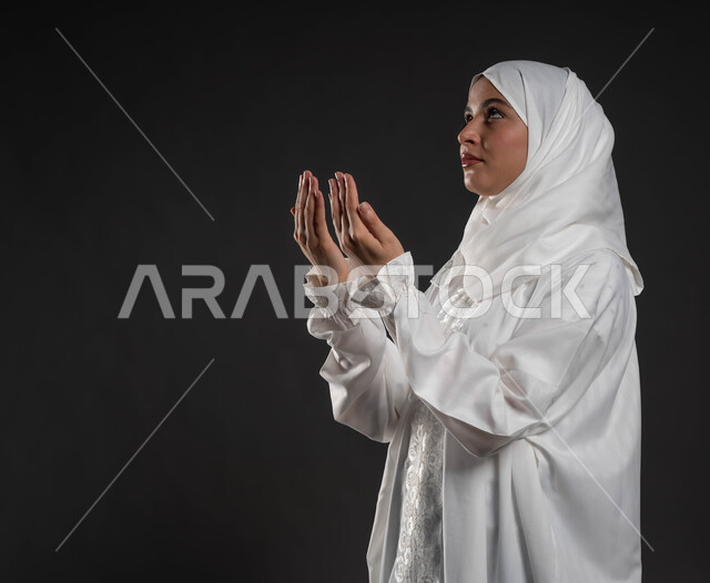 أداء الفرائض بأوقاتها، الدعاء والتضرع لرب العباد، العبادة والتقرب إلى الله، الركوع والسجود والخشوع، مفهوم أركان الإسلام، بورتريه مقرب لشابة عربية خليجية سعودية مسلمة ترتدي الحجاب و العباءة البيضاء تقف على السجادة، خلفية رمادية