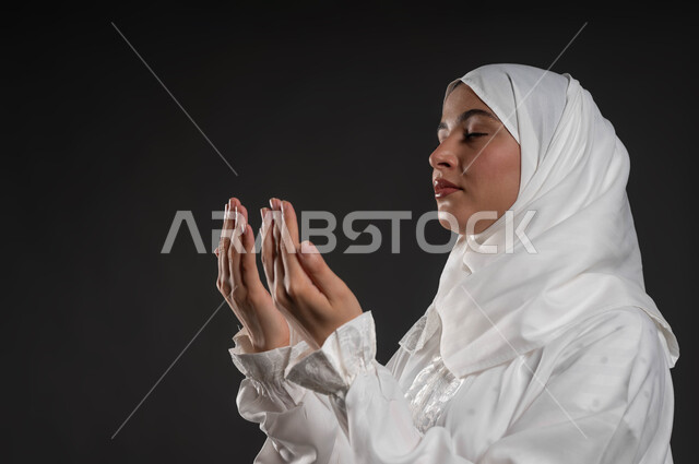 الدعاء والتضرع لرب العباد، العبادة والتقرب إلى الله، الركوع والسجود والخشوع، أداء الفرائض بأوقاتها، مفهوم أركان الإسلام، بورتريه مقرب لشابة عربية خليجية سعودية مسلمة ترتدي الحجاب و العباءة البيضاء تقف على السجادة، خلفية رمادية