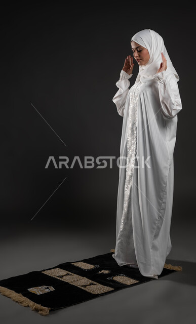 العبادة والتقرب إلى الله، الركوع والسجود والخشوع، الإقامة و الاستعداد للبدأ بالصلاة، أداء الفرائض بأوقاتها، مفهوم أركان الإسلام، بورتريه مقرب لشابة عربية خليجية سعودية مسلمة ترتدي الحجاب و العباءة البيضاء تقف على السجادة، صورة الجسم بالطول الكامل، خلفية رمادية
