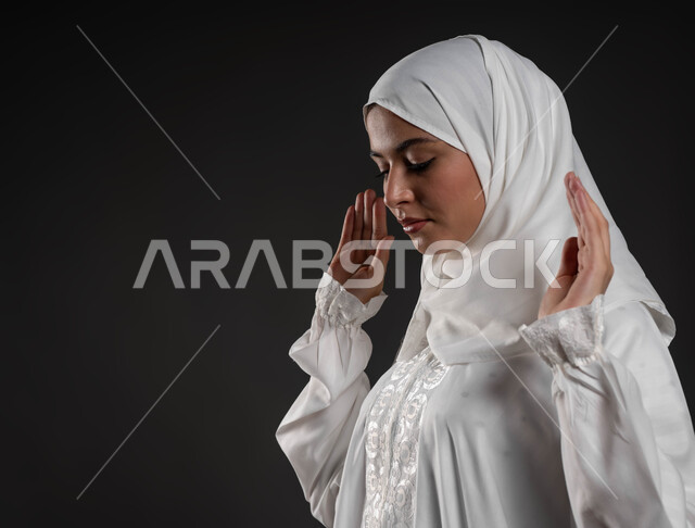 Staying and preparing to start praying, performing the obligatory prayers at their appointed times, the concept of the pillars of Islam, a close-up portrait of a young Arab Gulf Saudi Muslim woman wearing the hijab and white abaya, standing on the carpet, worship and getting closer to God, kneeling, prostrating, and reverence, gray background.