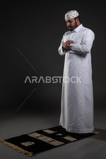 مفهوم أركان الإسلام ،أداء فرائض الصلاة بأوقاتها، بورتريه لرجل عربي خليجي سعودي مسلم يرتدي الثوب والقبعة الإسلامية البيضاء يقف على السجادة، العبادة والتقرب إلى الله، الركوع و السجود والخشوع، قراءة ما تيسر من القرآن الكريم، صورة الجسم بالطول الكامل ،خلفية رمادية